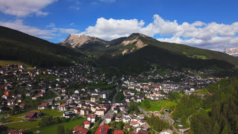 Drone footage of an alpine town surrounded by green hills and forested slopes, with a dramatic rocky mountain peak in the background in the Dolomites, Italy