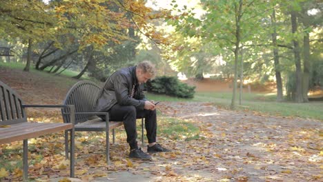 A-young-man-looking-at-his-mobile-phone-on-a-park-bench-alone-on-at-autumn-day