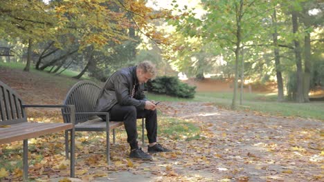 a young man looking at his mobile phone on a park bench alone on at autumn day