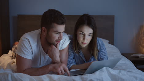 smiling young just-married couple scrolling and typing on the laptop while lying on the bed in the evening