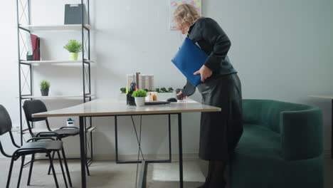 professional woman enters workspace, approaches desk, places file and coffee cup on surface, preparing to begin productive work session in modern minimalist office environment