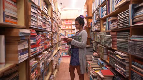 Asian-girl-standing-between-the-rows-of-bookshelves-and-exploring,-Side-angle-shot