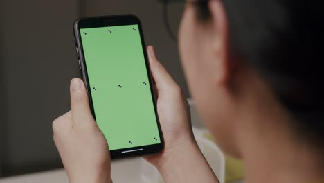 vista trasera de una mujer caucásica sosteniendo un teléfono inteligente de pantalla verde con llave de croma viendo contenido en línea. mujer usando teléfono móvil, navegando por internet, viendo contenidos en casa.