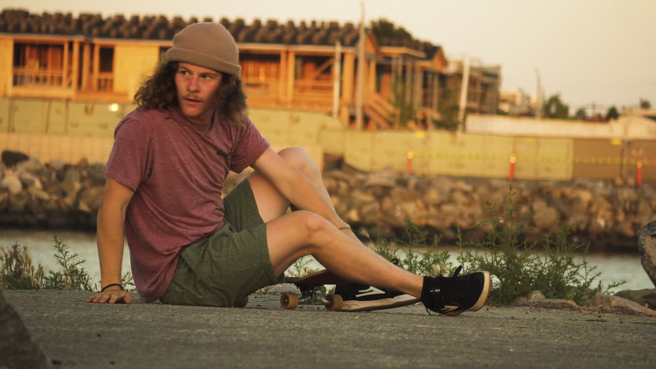 Skateboarder with long curly hair gets up from ground, puts his beanie on and rides away