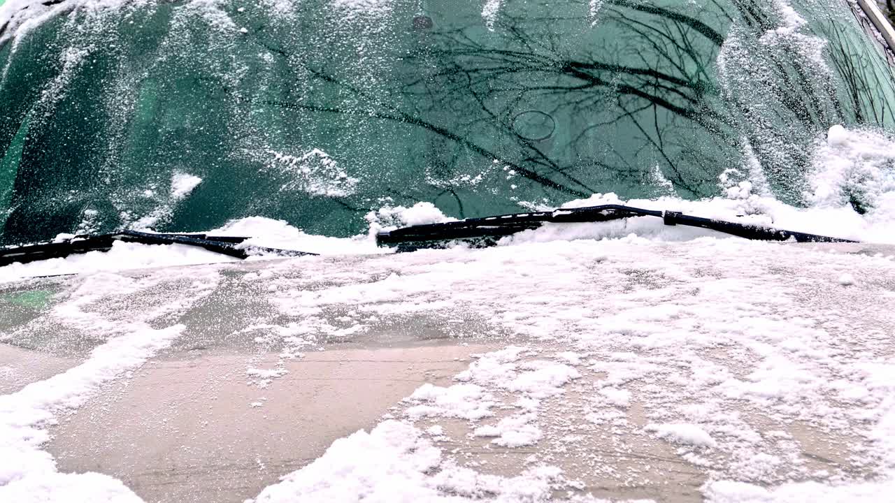 A person uses a blue snow brush to clear heavy snow from a car's windshield on a winter morning