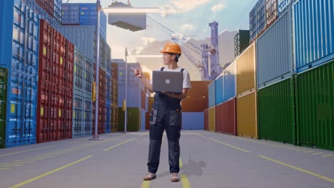 cuerpo lleno de hombre asiático trabajador con gafas de protección y casco de seguridad usando una computadora portátil y apuntando a un lado mientras estaba de pie en el almacén del patio de contenedores