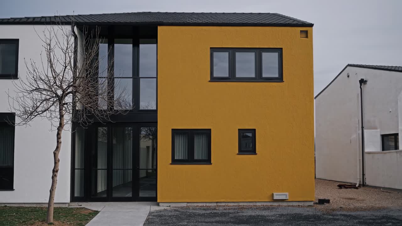Contemporary yellow residential building with black roof and windows, showcasing modern architectural design in a residential neighborhood, featuring a bare tree and a paved pathway