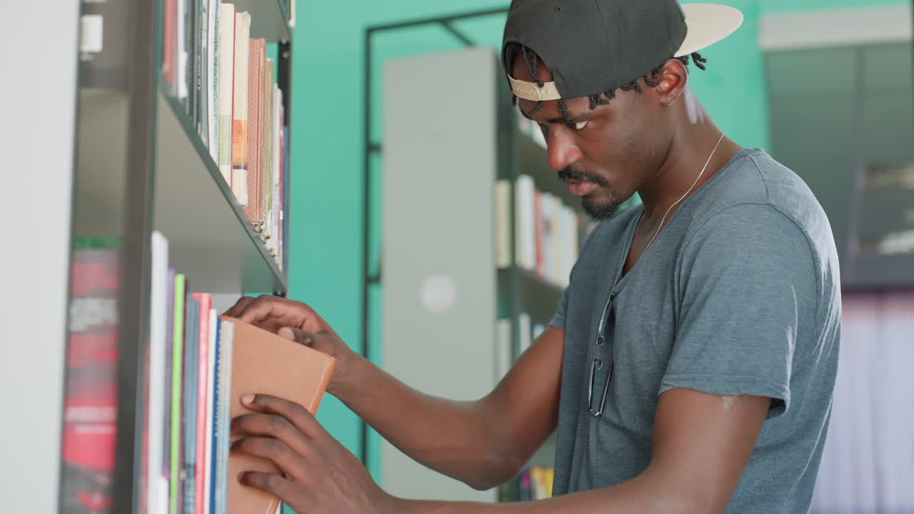 Sidebillede af ung mand i fritidstøj, der sætter en brun bog tilbage på bibliotekets hylde. Han har kasketten bagvendt på, et fokuseret udtryk og står mellem rækker af bøger i rolige akademiske omgivelser