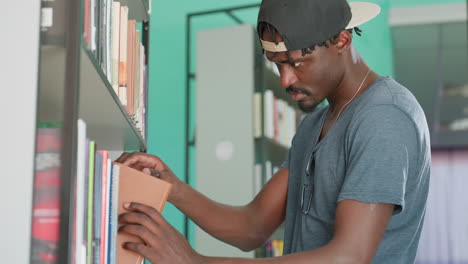 Side view of young man in casual clothing returning brown cover book to library shelf, wearing cap backward, focused expression, standing among rows of books in quiet academic environment