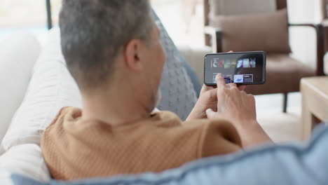 happy senior biracial man using smartphone on couch in sunny living room, slow motion