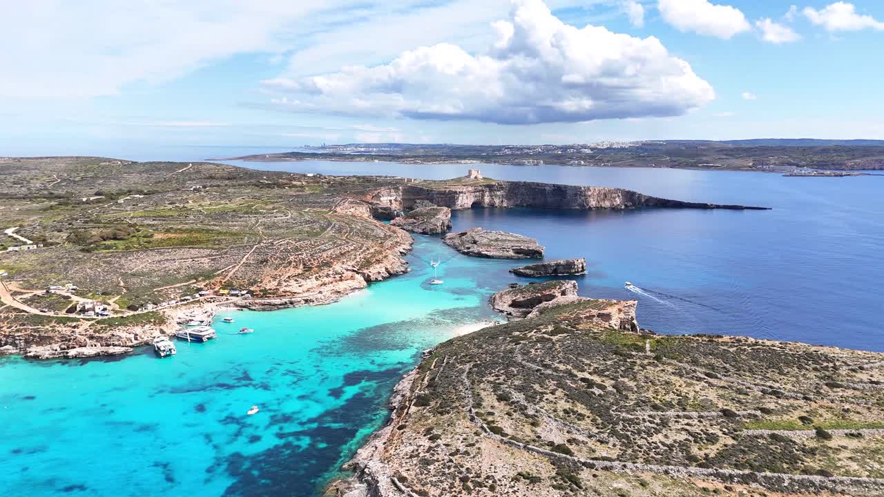 Droneutsikt over Den blå lagune i Comino, Malta, med flere båter ankret og i bevegelse over grunt turkist vann, innrammet av steinete kystlinje og åpent hav.