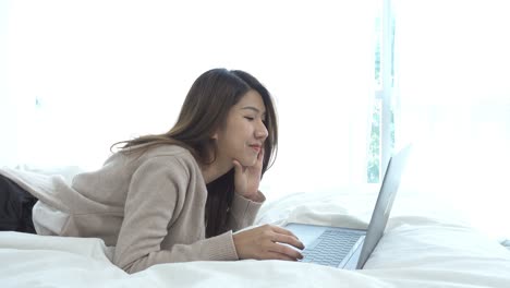 아름다운 미소 짓는 젊은 아시아 여성은 집에서 침실에서 침대에 앉아 노트북에서 일합니다.