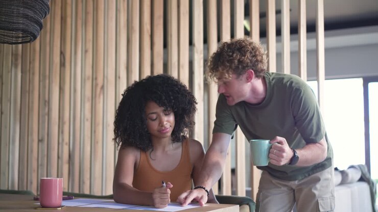Diverse couple reviewing papers at wood table as man leaning with mint mug, guiding pen, embracing