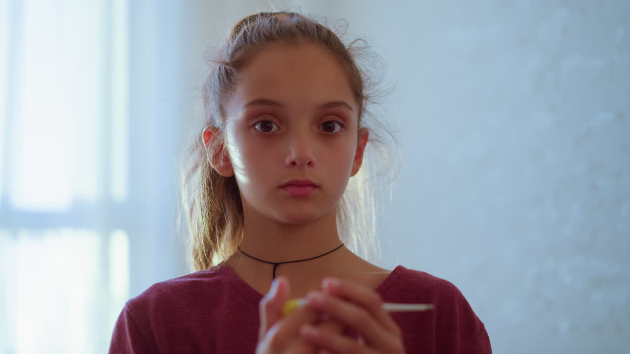 Thoughtful girl closely examines thermometer with calm serious expression showing concern care and awareness in softly lit indoor environment suggesting emotional responsibility