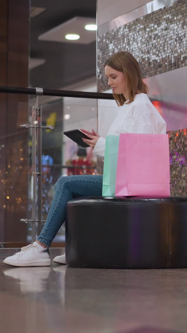 joven sentada en un banco en un centro comercial, deslizando a través de su teléfono con bolsas de compras de colores a su lado y decoraciones festivas, alguien es visible en el fondo en el ascensor