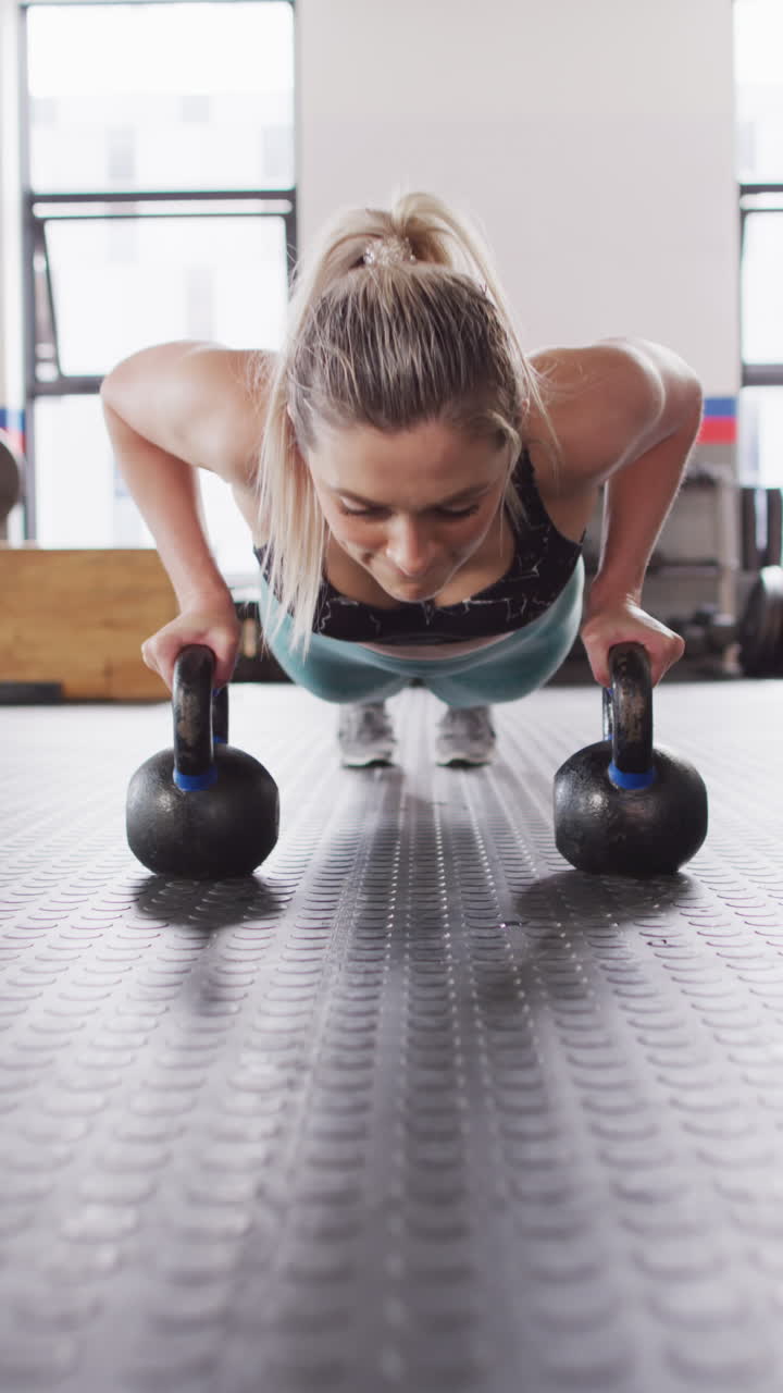 Vertical video of determined caucasian woman doing push ups on kettlebells working out at gym