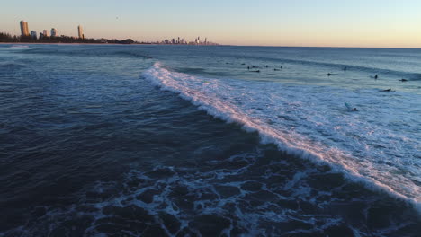 Luftaufnahmen-Von-Drohnen,-Die-Surfer-Entlang-Brechender-Wellen-Verfolgen,-Mit-Oberflächenparadies-Qld-Im-Hintergrund-Während-Des-Sonnenaufgangs-Am-Beliebten-Touristenziel-Burleigh-Heads-Gold-Coast-Qld-Australien