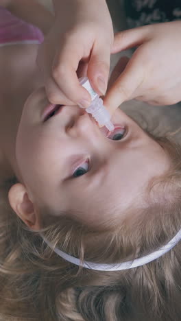 careful mommy gives eyes drops to smiling little girl lying on large comfortable bed close upper view