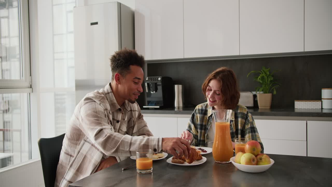 joven hombre moreno negro feliz tomando el desayuno con su joven novia adulta con cabello marrón en un peinado de bob en una camisa a cuadros verde en la mesa de la cocina por la mañana en un apartamento moderno
