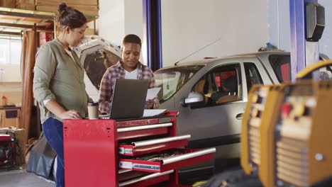 Video-of-two-diverse-female-car-mechanics-using-laptop-and-smartphone,-searching-for-car-parts