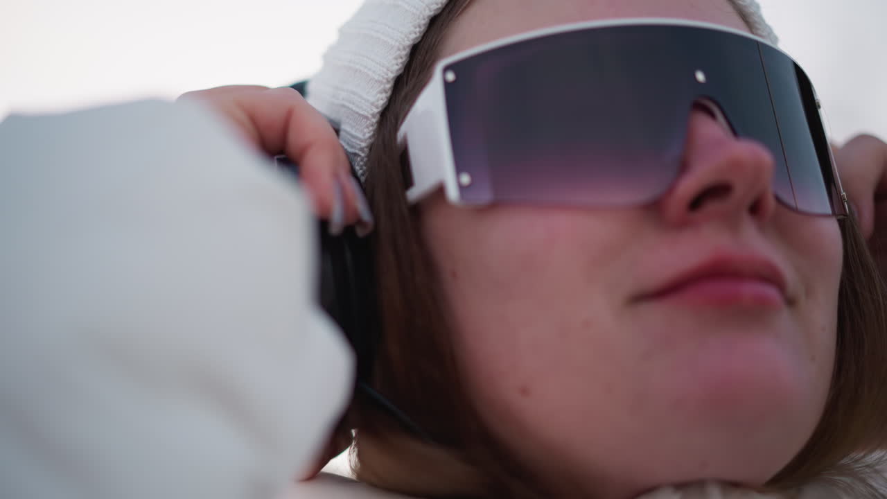 Close up of young artist wearing over ear headphones smiling while listening to music against blurred winter background snow glistens under sun while she dons beanie and coat enjoying breeze