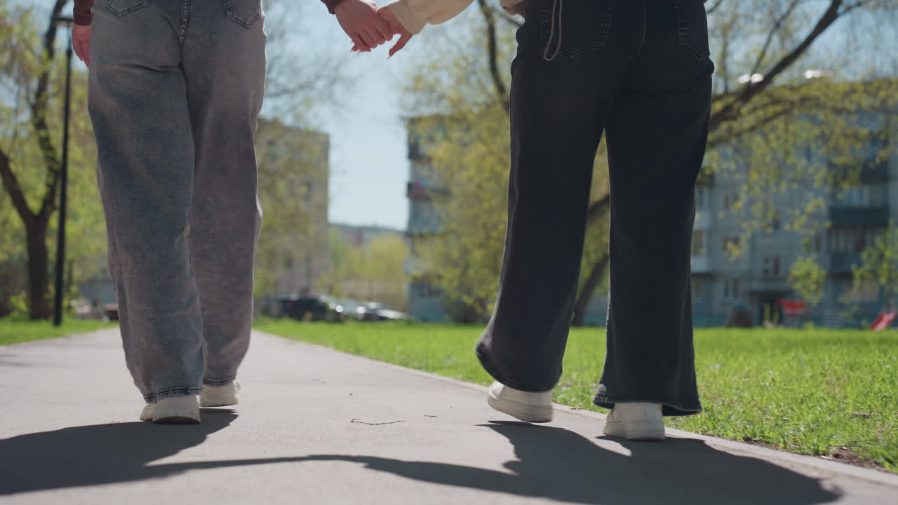 백인 여성과 아시아 여성이 나무 사이 그늘진 길을 따라 걸어가고 있고, 따뜻한 역광과 얼룩덜룩한 햇빛이 비치며, 편안한 속도와 부드러운 동료애가 느껴지고, 녹지 공간 너머로 교외 건물들이 보입니다.