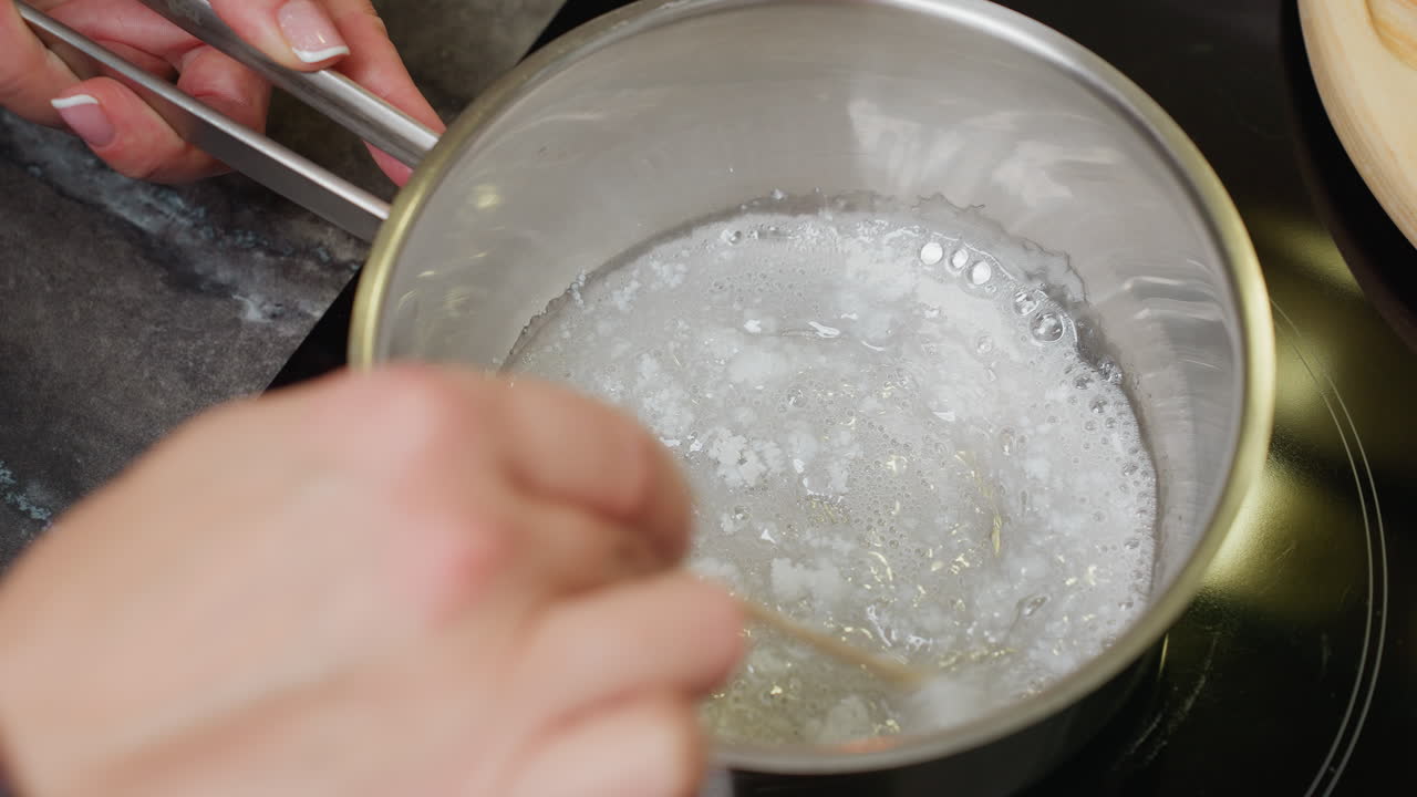 Primo piano della mano di una persona che mescola lo zucchero in una pentola, processo di cottura, zucchero che si scioglie e forma uno sciroppo, azione in una pentola sul piano cottura, scena in cucina, attenzione alla cottura dello zucchero per dessert o dolci