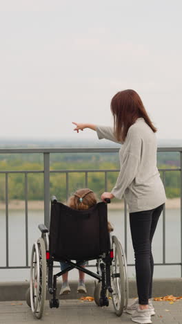 woman shows large park with green trees to daughter with spinal cord injury. mother and little girl in wheelchair on observation deck overlooking river