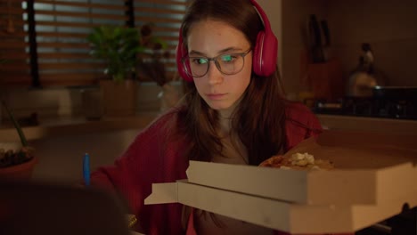 confident brunette girl in red wireless headphones with glasses studying foreign languages and learning new things while making a snack and eating pizza from a packaging box in a modern apartment in the kitchen