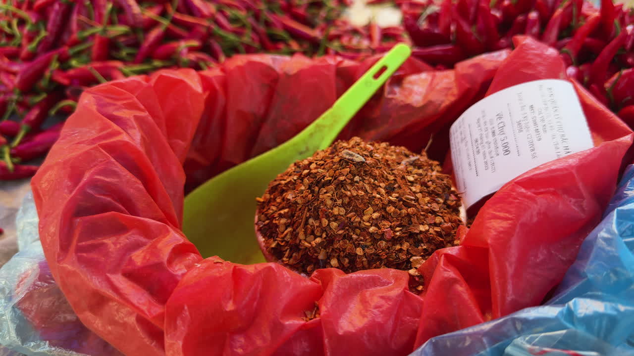 Primer plano de condimento de chile triturado en una bolsa de plástico roja con cucharón y etiqueta, rodeado de chiles rojos frescos en un puesto de mercado de alimentos al aire libre.