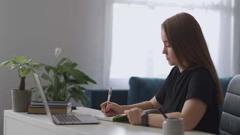 Studentin-Sieht-Sich-Vorlesungen-über-Das-Internet-An-Und-Macht-Sich-Notizen-In-Ihr-Notizbuch,-Sitzt-Mit-Laptop-Am-Tisch-Und-Schaut-Auf-Das-Display,-Lernt-Von-Zu-Hause-Aus-Fernunterricht