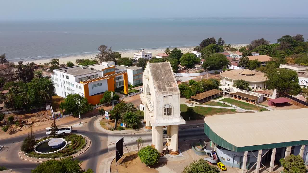 Aerial parallax shot of the Arch 22 memorial the triumphal arch of banjul city in the gambia with a view of the sea and a busy road with traffic at a roundabout and buildings on a beautiful beach