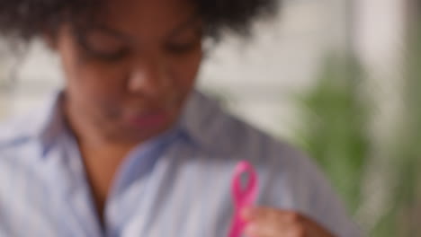 Primer-Plano-De-Una-Mujer-Adulta-Sonriente-En-Una-Clínica-Hospitalaria-Sosteniendo-Una-Cinta-Rosa-De-Concientización-Sobre-El-Cáncer-De-Mama-Y-Colocándola-En-Su-Camisa-1