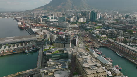 Hacia-Atrás-Volar-Por-Encima-De-Los-Edificios-En-El-Puerto.-Varios-Barcos-Parados-En-Los-Muelles.-Inclinar-Hacia-Arriba-Revela-El-Paisaje-Urbano-Y-La-Majestuosa-Montaña-De-La-Mesa.-Ciudad-Del-Cabo,-Sudáfrica