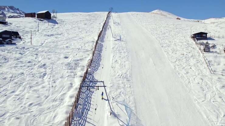 Aerial view following snowy ski resort drag lift carrying winter tourists up steep mountain slope in Farellones, Santiago, Chile