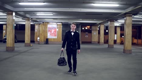 Young-blonde-man-in-a-black-suit-with-bow-tie-walking-on-the-parking-with-big-leather-bag.-Slow-Motion