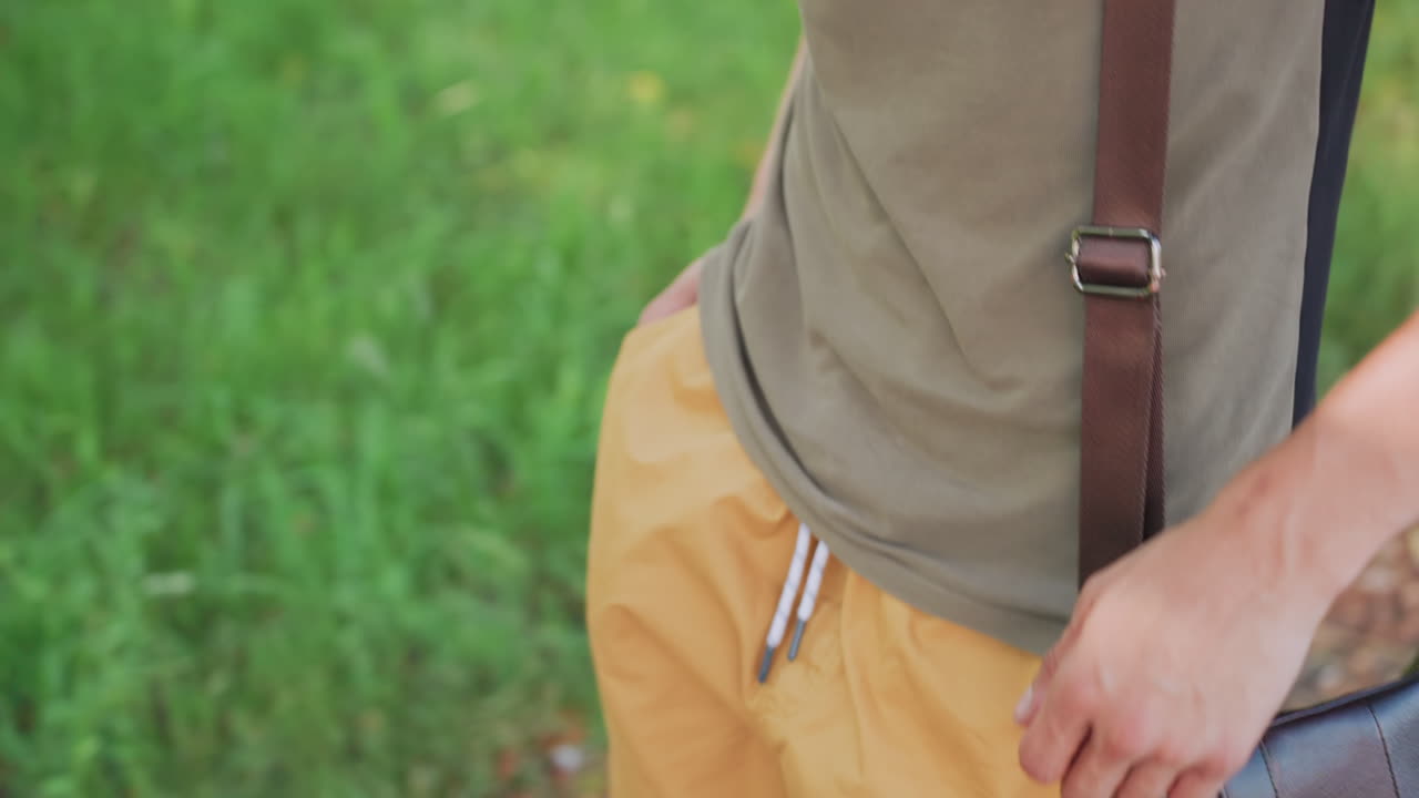 Young Person Handling Fabric Carefully, Young Man Performing Small Grooming Ritual On Outdoor Grass, Young Adult Meticulously Wiping Bag Strap With Tissue Amidst Shaded Grassy Environment