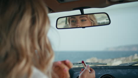 woman looking car mirror correcting hairstyle close up. blonde holding lipstick.