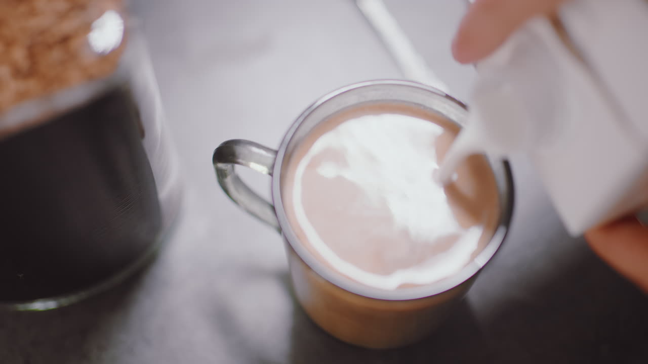 Close up of fresh dairy milk pouring into steaming coffee inside glass mug, creating smooth swirl and creamy texture, highlighting warm morning beverage preparation