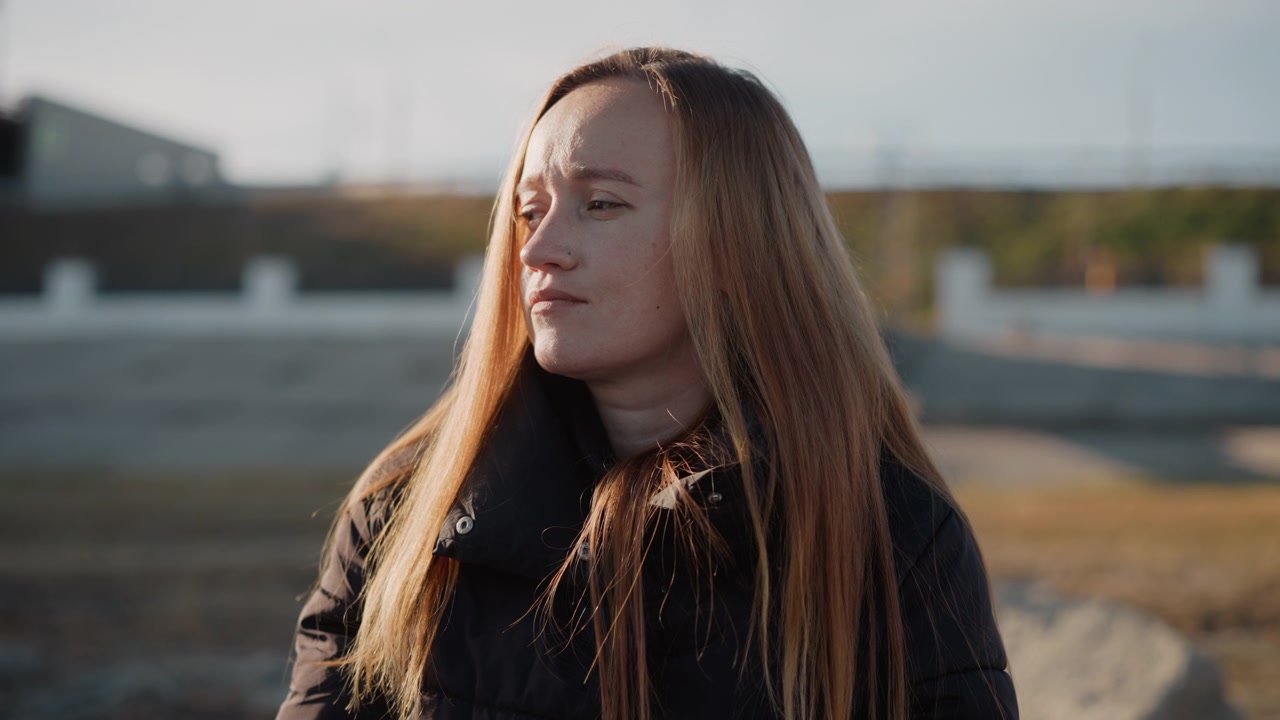 Kvinde med langt hår står udendørs i afslappede omgivelser iført mørkt tøj. Baggrunden viser et åbent område med blødt naturligt lys og fjerne strukturer, der antyder dagslys. Hendes holdning.