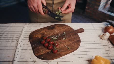 a person chopping fresh rosemary next to cherry tomatoes on a wooden board