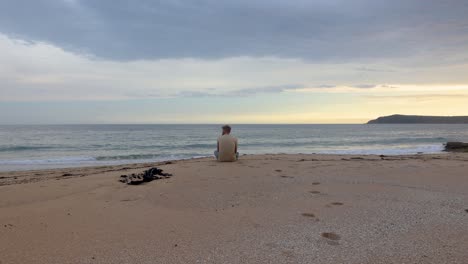 a man sits on the shoreline of a beautiful beach on the australian coast and looks out to sea at the sunset