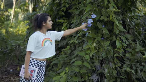 Ein-Wunderschönes-Indisches-Mädchen-Mit-Weißem-Oberteil-Und-Einem-Buch-In-Der-Hand,-Das-Eine-Blume-Von-Der-Pflanze-Pflückt