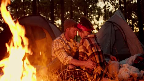 Romantic-evening,-a-man-in-a-checkered-shirt-and-a-hat-sits-near-a-girl-with-a-red-bandana-and-in-a-checkered-shirt-near-a-fire-against-the-backdrop-of-tents-and-they-closely-communicate-with-each-other-about-something-against-of-a-forest-in-the-fall