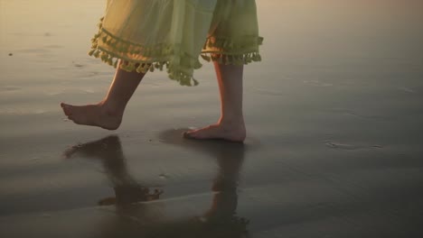 Feet-of-a-woman-in-a-dress-walking-at-the-shoreline-on-wet-sand-with-her-shadow-showing-as-the-sun-setting