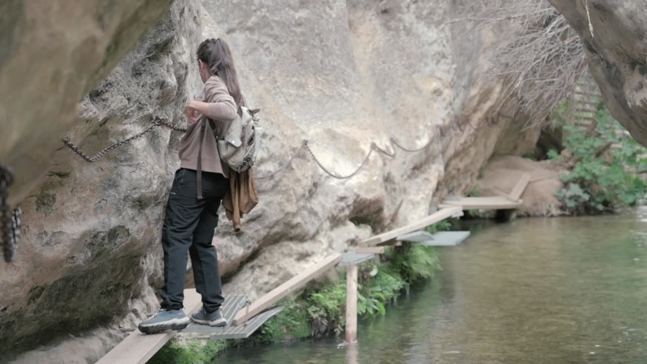 Person walking along a narrow wooden boardwalk attached to a rocky cliffside, using a safety chain for support