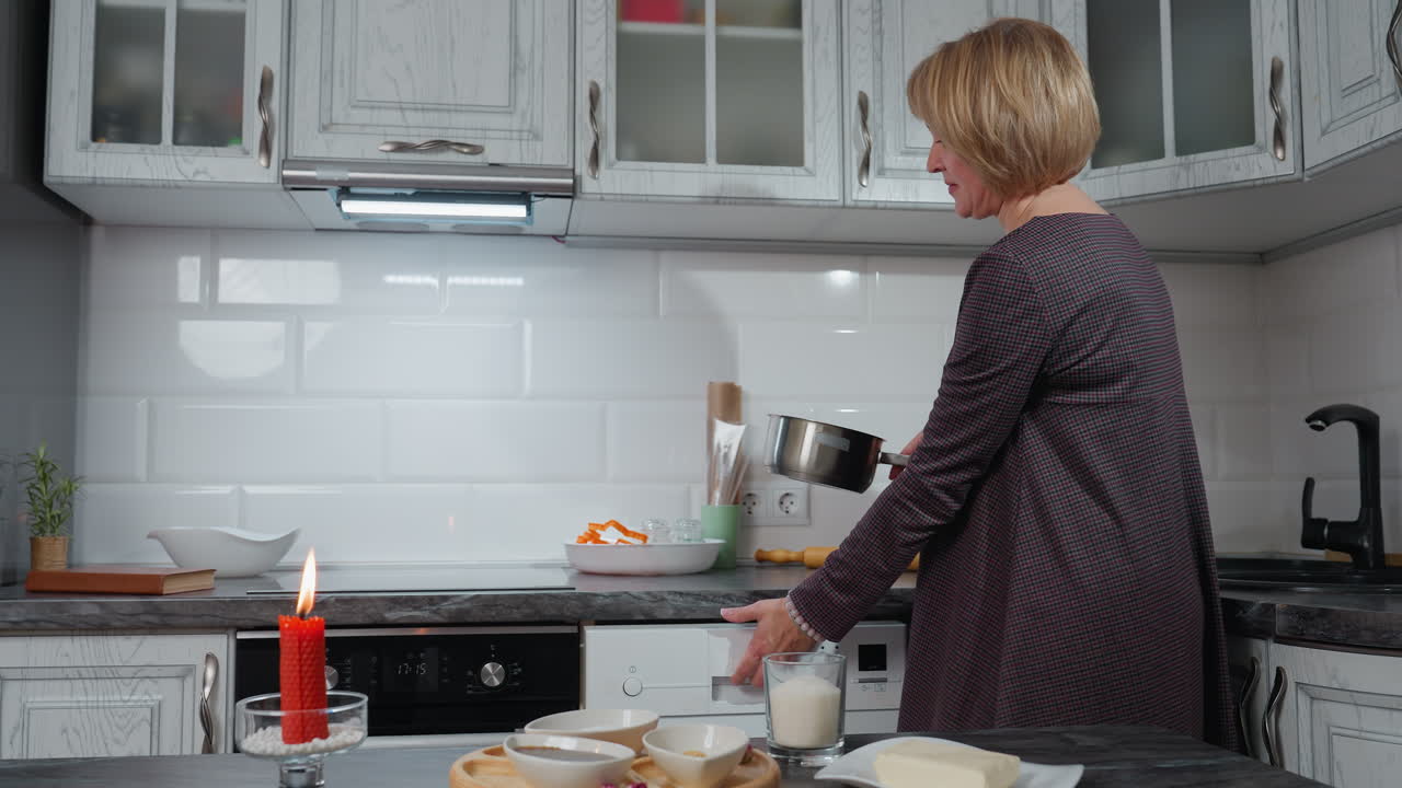 Woman in elegant gown bends to retrieve pot from lower kitchen cabinet while table is set with a burning red candle, sugar, butter, and spices