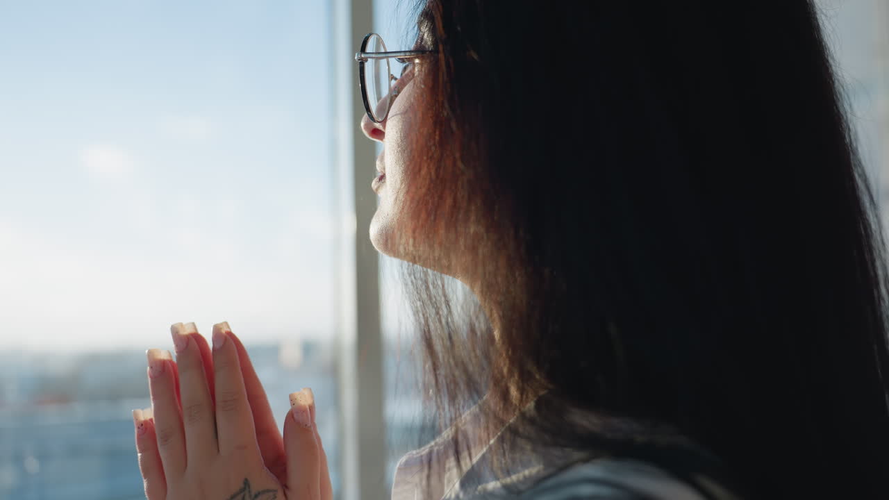 Close up of lady calmly pausing with book balanced on head while gazing through building glass wall, bathed in warm sunlight streaming into modern space
