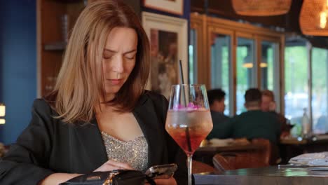 a stylish woman is intently engaged with her smartphone while she enjoys sipping a cocktail at a modern bar