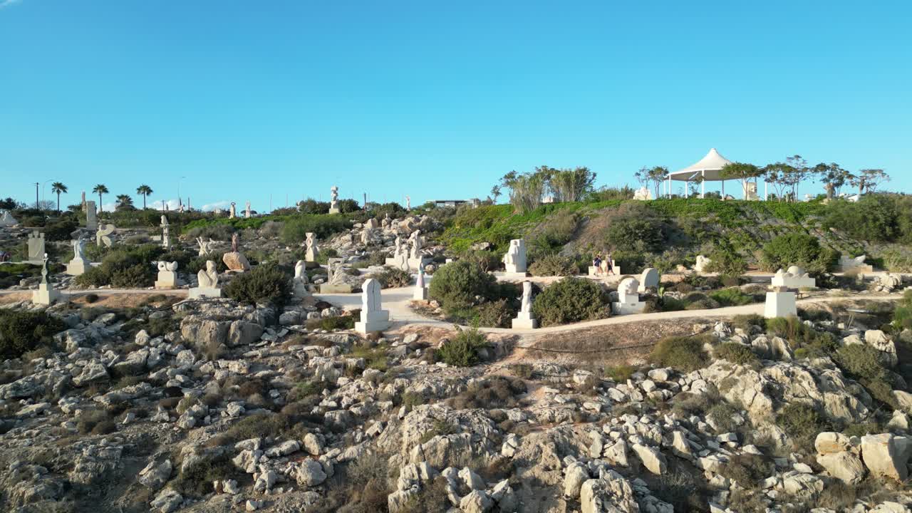 Drone tracking along walking path of Ayia Napa Sculpture Park showcasing unique stone art near the sea cliffs under blue sky